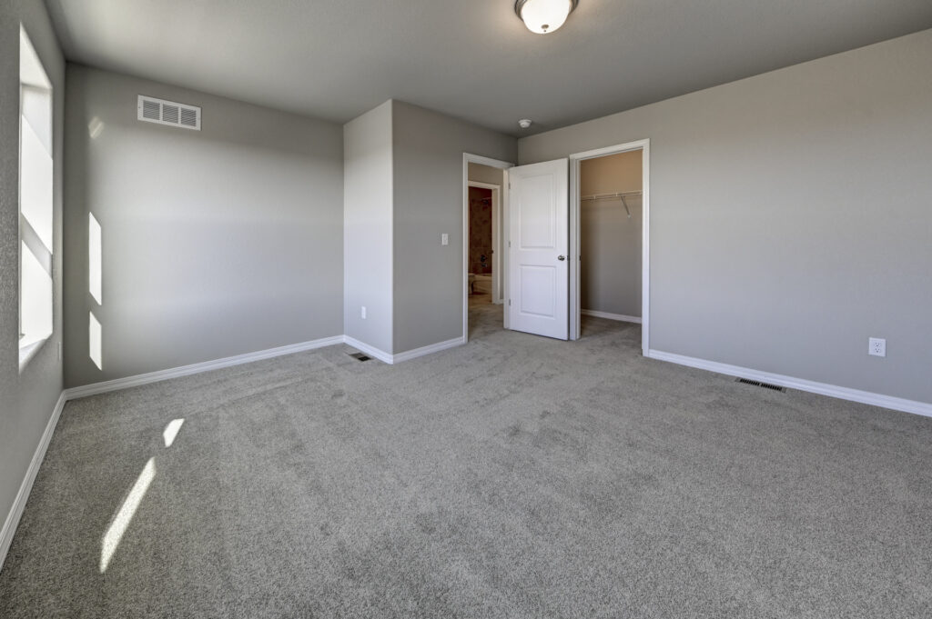Large carpeted bedroom with open door to hallway and closet, two windows letting in sunlight.