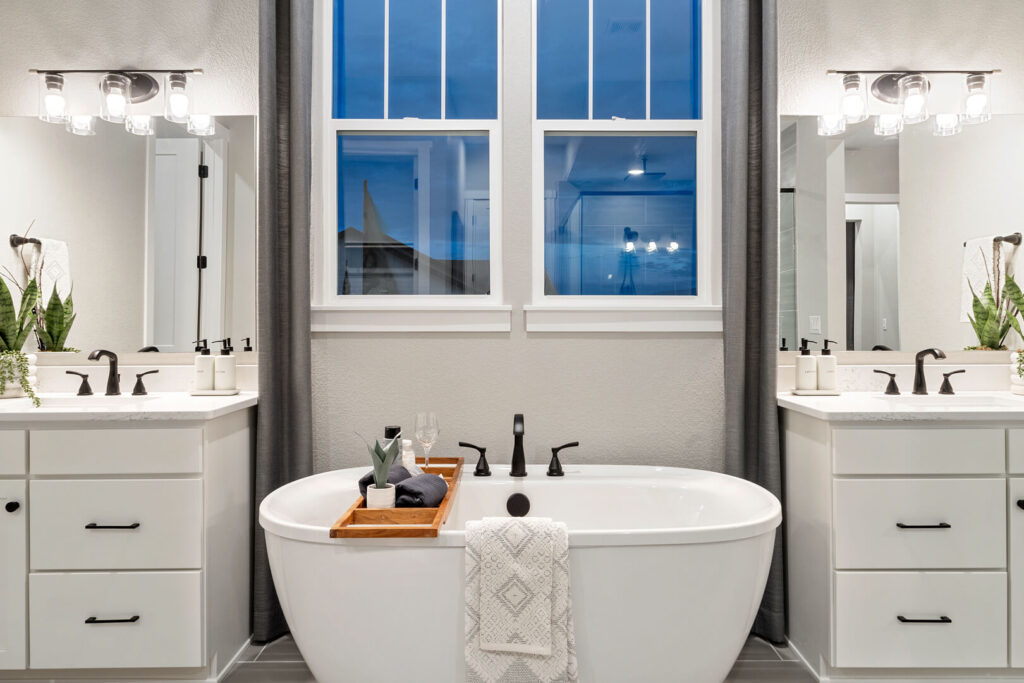 Close view of a freestanding bathtub with wooden tray, twin vanities, and a double window.
