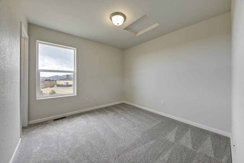 Empty carpeted bedroom with single window and ceiling light, neutral walls and baseboards.