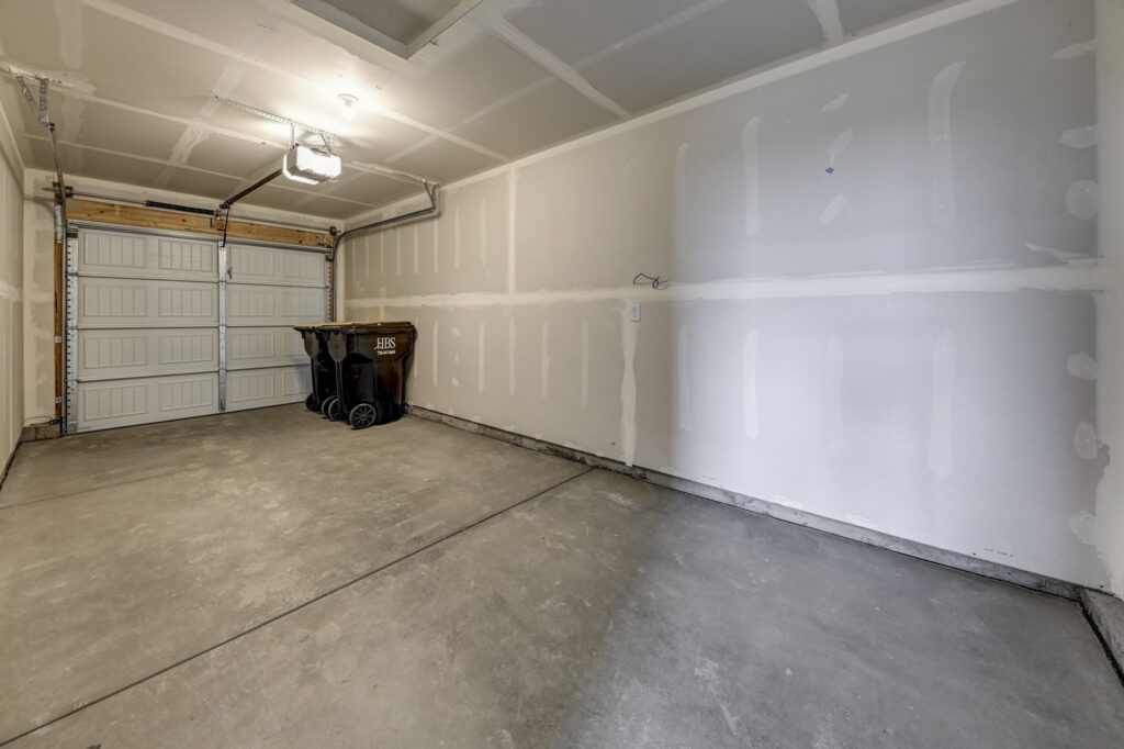 Empty single-car garage with unfinished drywall and concrete floor, two trash bins at rear.