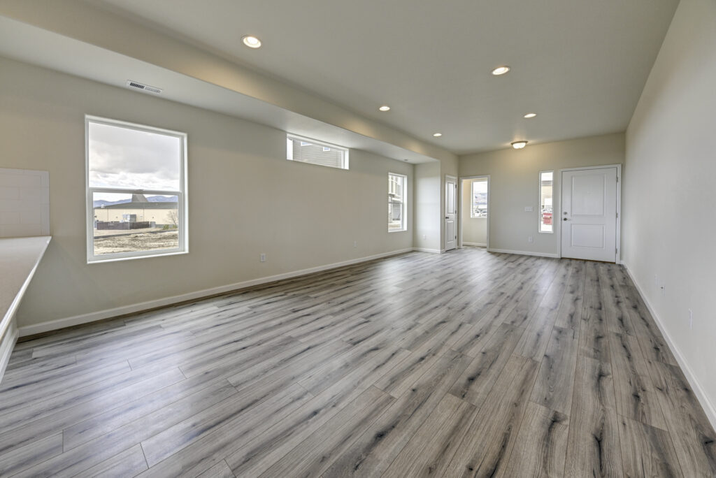 Wide empty living area with light wood-look flooring and several windows under recessed ceiling lights.