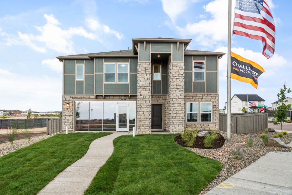 Two-story model home with stone facade, front lawn, and flags on a flagpole.