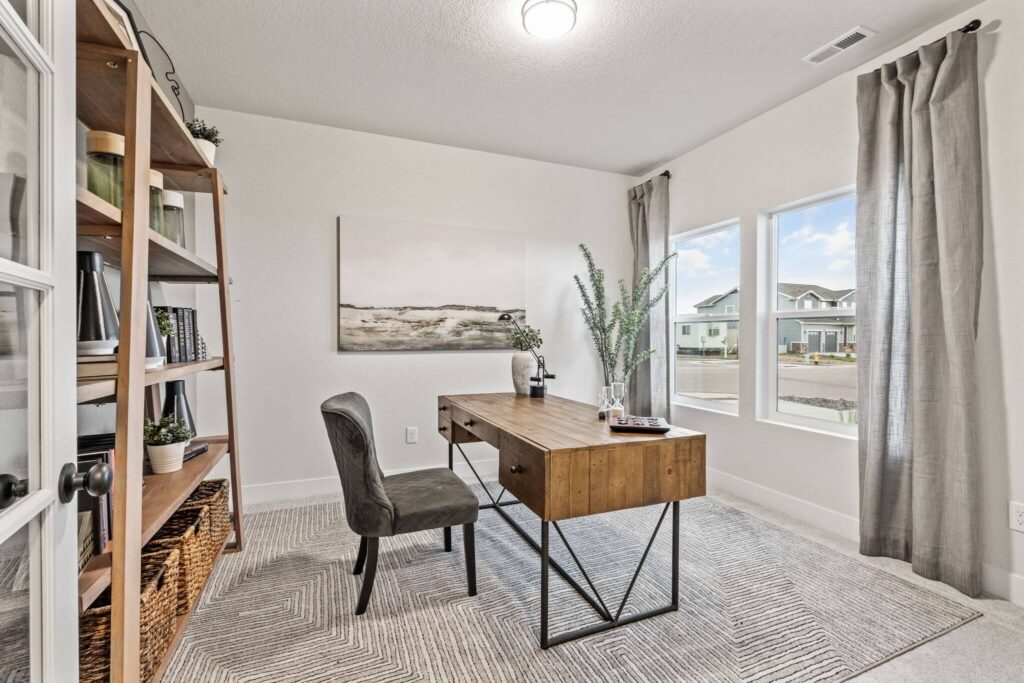 Home office with wooden desk, gray chair, open bookshelf and three front windows.
