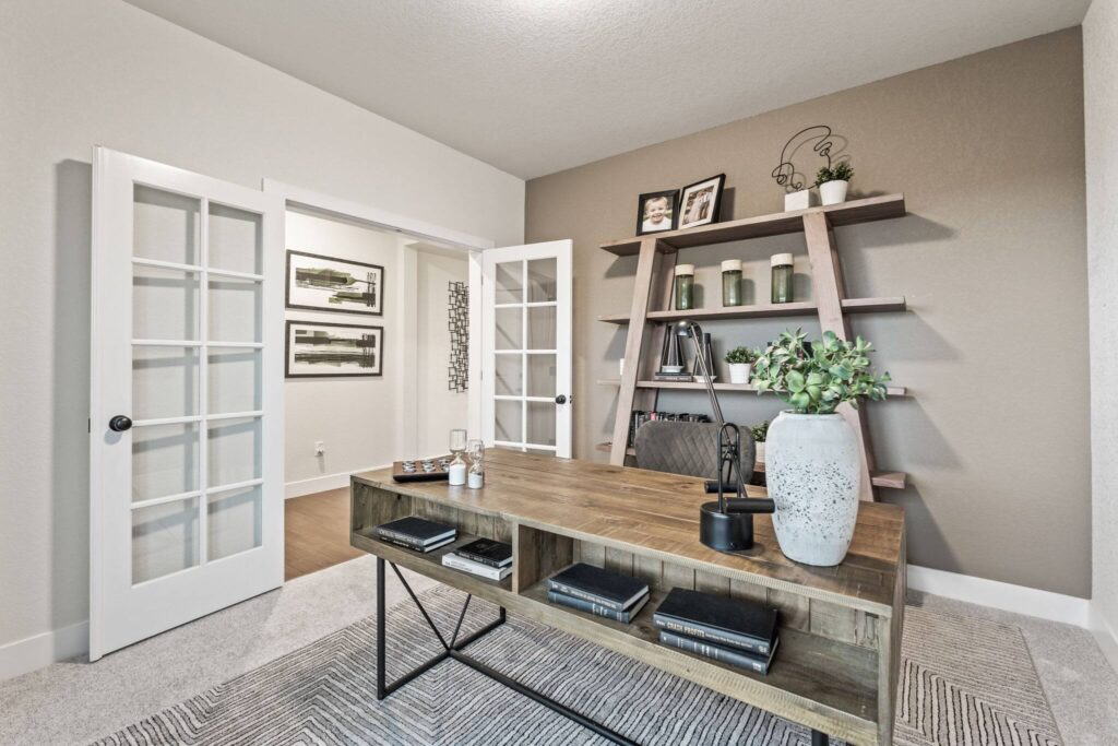 Office interior with wooden desk, shelving unit against taupe accent wall, and open French doors.