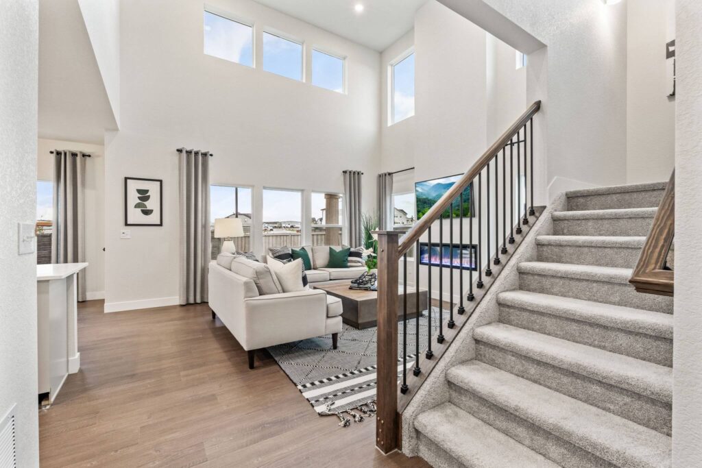 Two-story living room with carpeted staircase, light sofas, and tall windows above a hardwood floor.