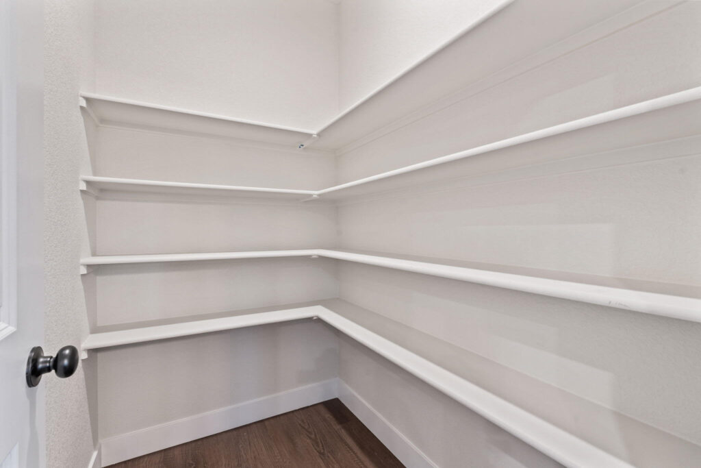 Small walk-in pantry with multiple white corner and wall-mounted shelves and wood-look flooring.