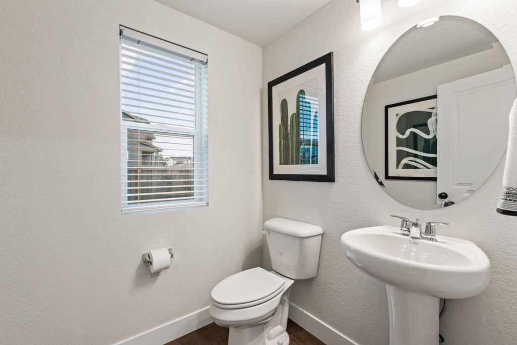 Small powder room with pedestal sink, toilet, round mirror, and framed artwork beside a window.