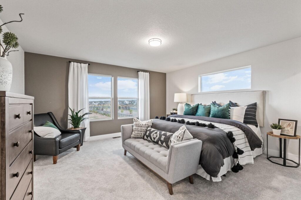 Staged bedroom with a bed, bench, armchair and two windows showing an outdoor view.