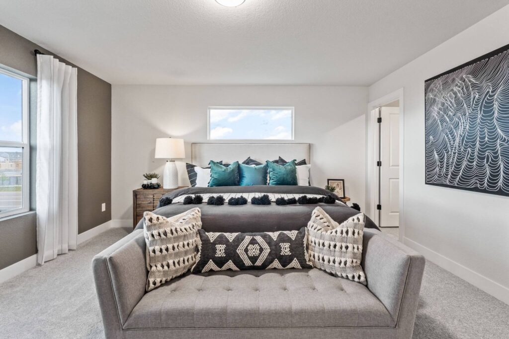Bedroom seen from the foot of the bed with a small loveseat, throw pillows and a window above the headboard.