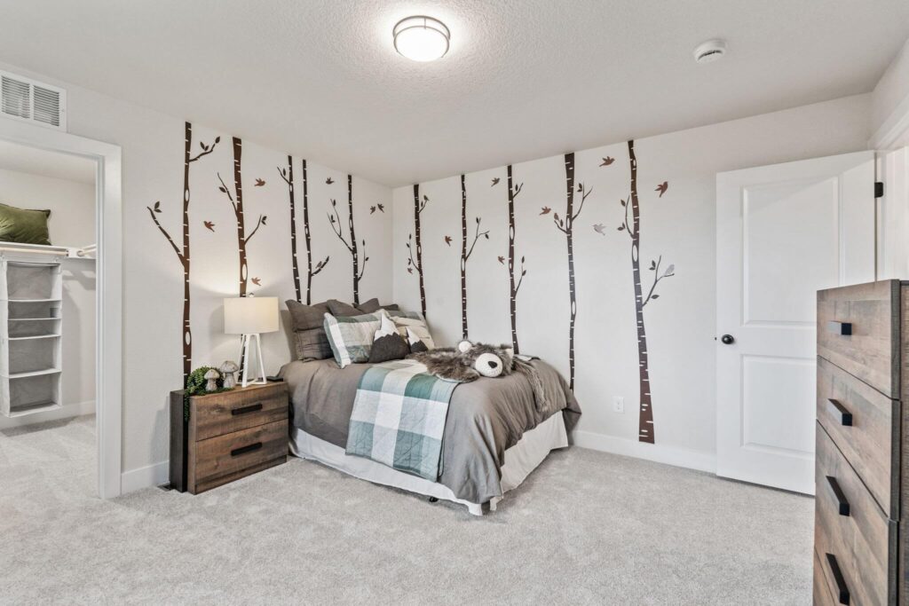 Staged bedroom featuring bed against a wall of tree decals and a nightstand with lamp.