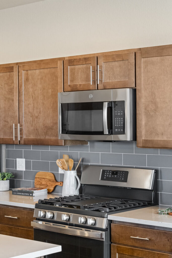 Close-up of kitchen stove area with stainless steel microwave, gas range, brown cabinets, and gray tile backsplash.