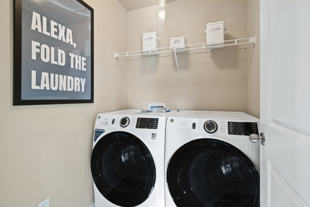 Compact laundry closet with front-loading washer and dryer under a wire shelf and wall art.