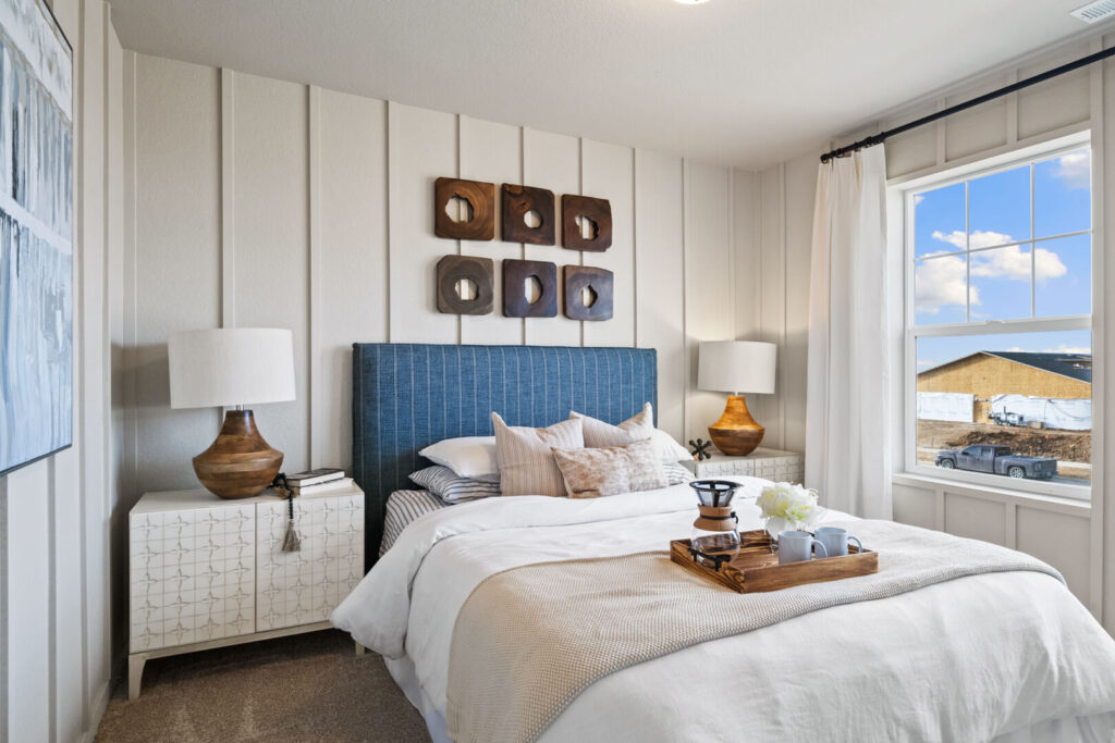 Staged bedroom with blue upholstered headboard, decorative wall art, and window showing exterior construction.