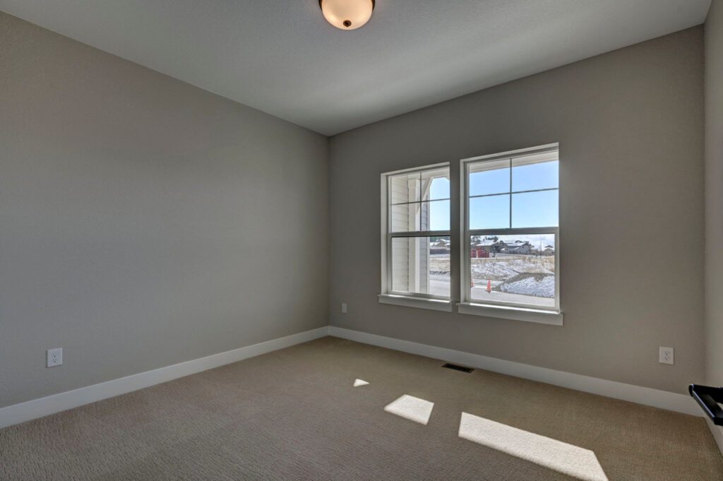 Empty carpeted room with two windows, beige walls, and a ceiling light fixture.