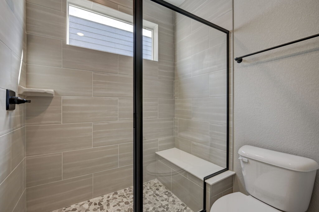 Walk-in shower with bench, horizontal light-gray wall tiles, and a high narrow window.