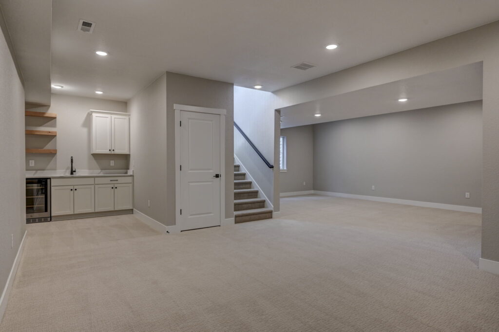 Open basement space showing wet bar, storage door, carpeted floors and stairwell to upper level.
