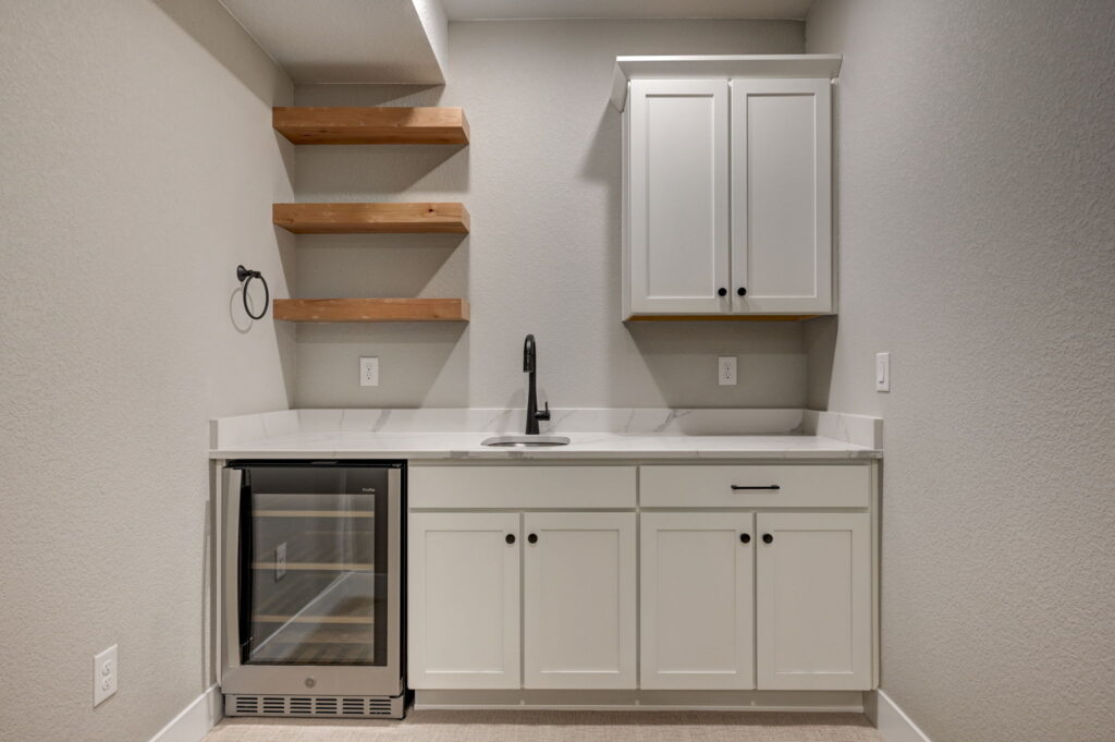 Small wet bar area with white cabinets, quartz countertop, built-in beverage fridge and open wood shelves.