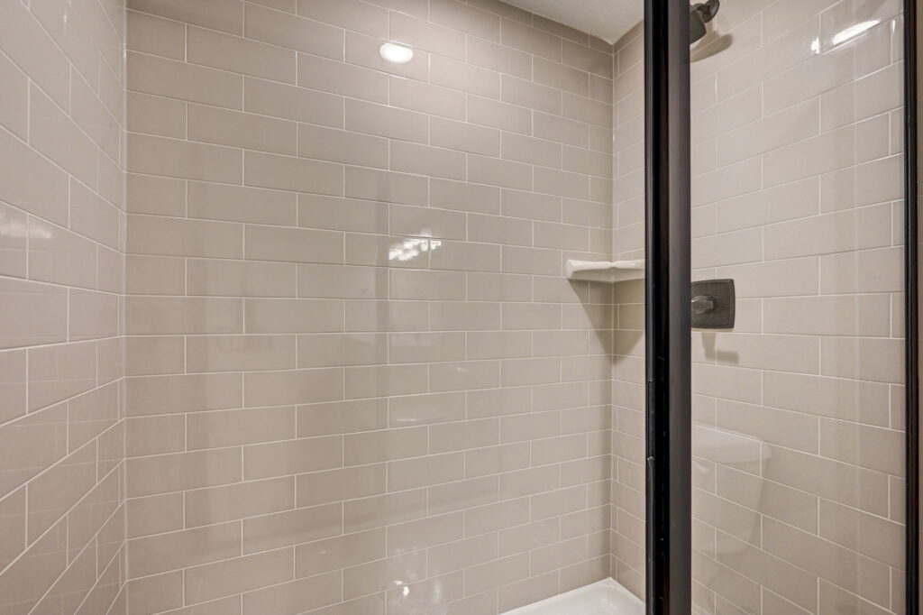 Frameless glass shower with light gray subway tile and small corner shelf.