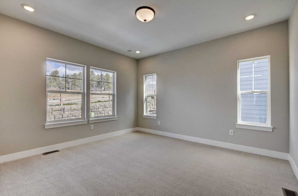 Empty carpeted room with three windows and beige-gray walls, recessed and ceiling light.