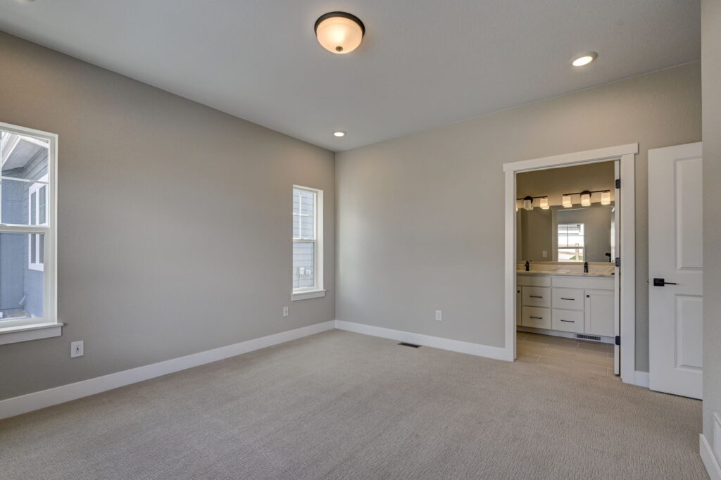 Empty bedroom showing two windows and an open doorway leading to a bathroom with vanity lights.