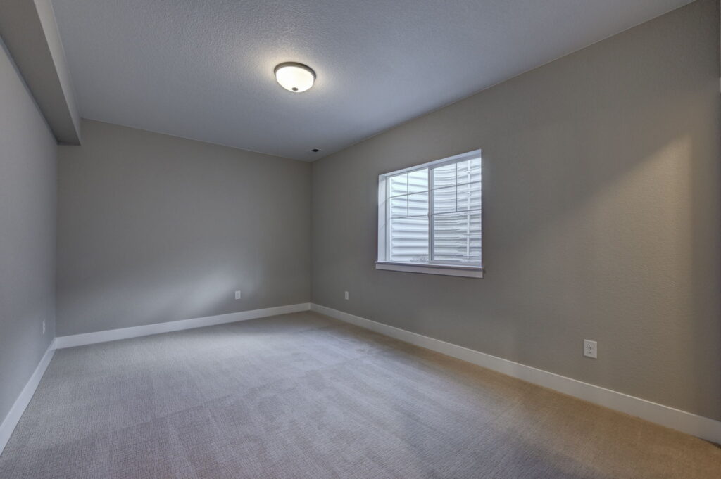 Empty carpeted room with a single window and neutral gray walls under ceiling light.