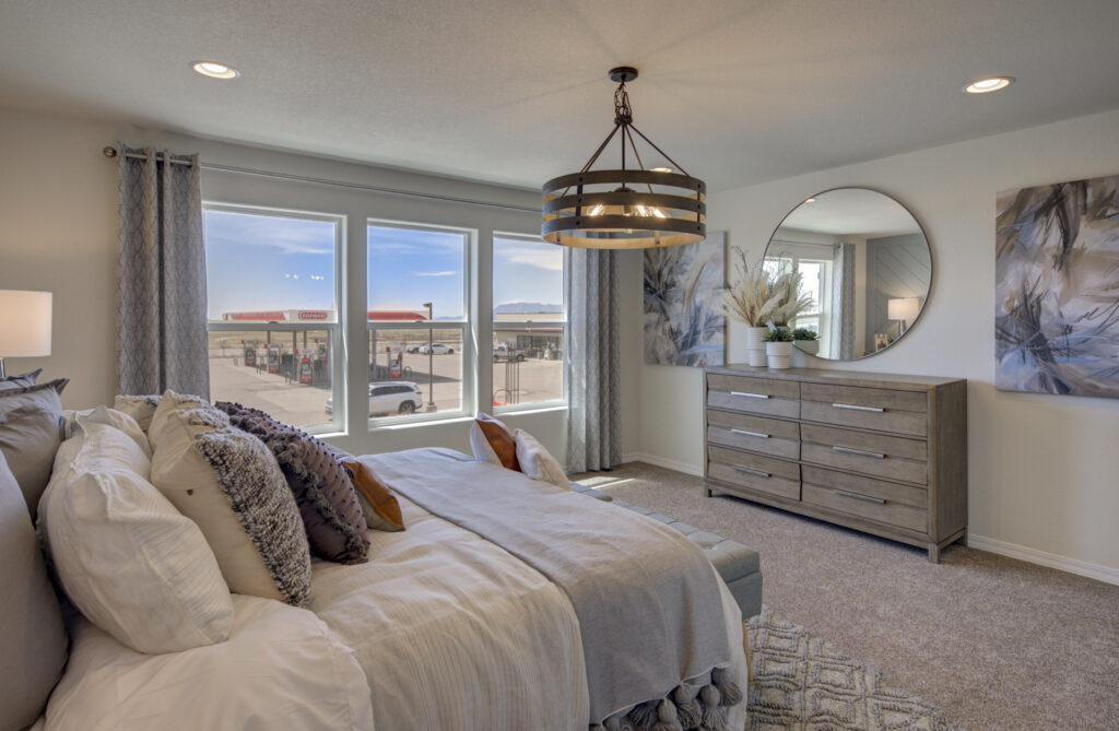 Bedroom interior with large front windows, dresser, round mirror and layered bedding.