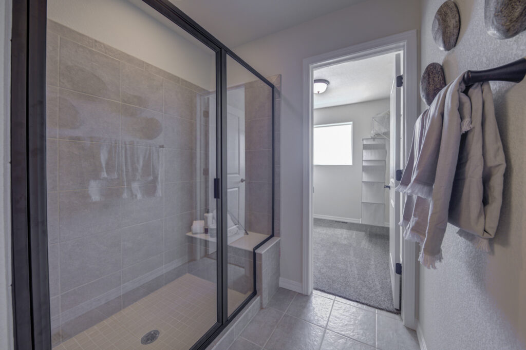 Glass-enclosed tiled shower with bench next to doorway leading to a small carpeted room.