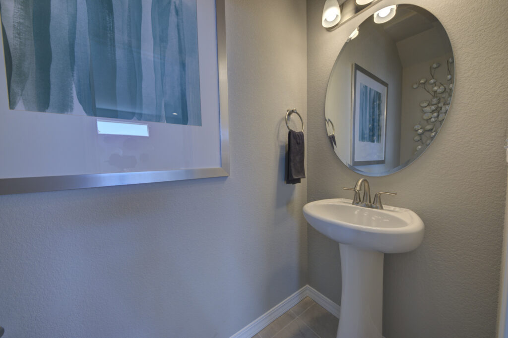 Small powder room with pedestal sink, round mirror, wall sconce, and framed artwork reflected in the mirror.