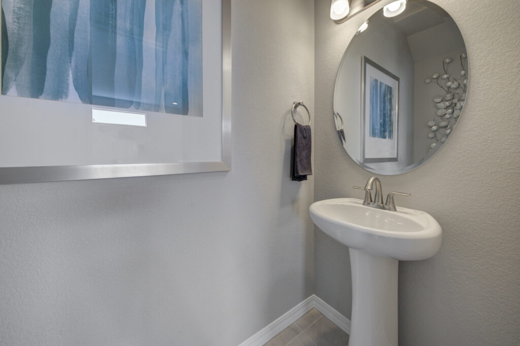 Narrow bathroom corner with pedestal sink, round mirror, and framed teal-striped artwork on the wall.