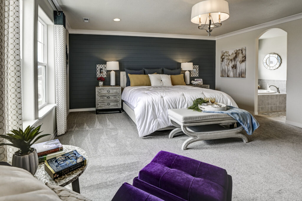 Wide bedroom view showing bed against dark accent wall, ottoman bench and doorway to bathroom.