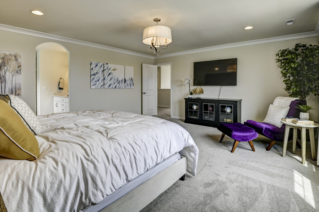 Bedroom interior with mounted TV, purple lounge chair and ottoman, and doorway to en suite.