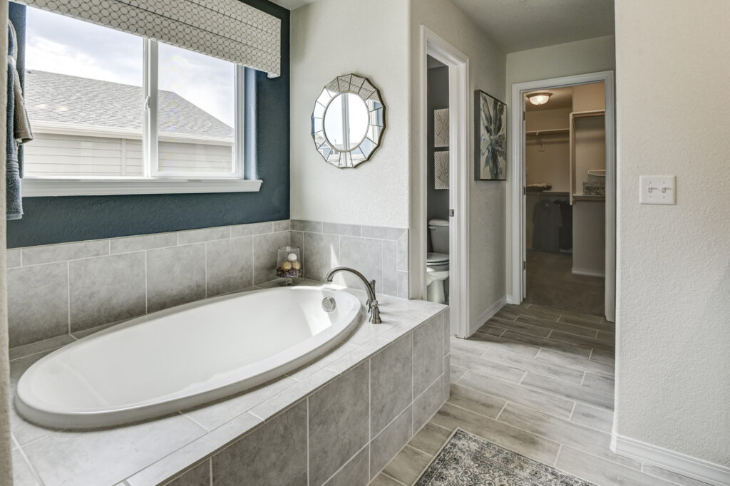 Built-in tiled bathtub under a window with a decorative round mirror and navy accent wall.