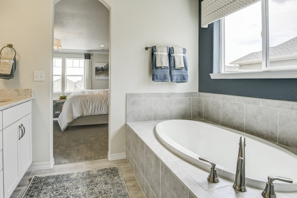 Edge of a tiled bathtub with towels on a rack and an arched opening to a bedroom.