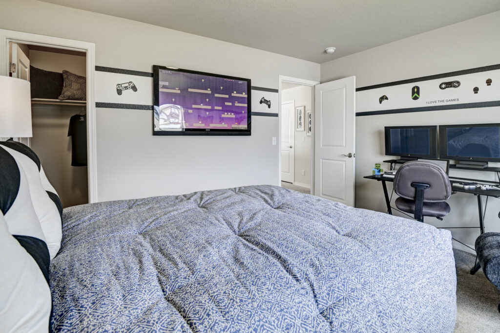 Bedroom view from foot of bed showing mounted flat-screen TV and dual computer monitors on desk.