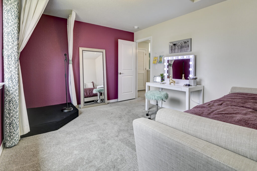 Bright bedroom with mauve accent wall, carpeted floor, vanity mirror, and small stage area.
