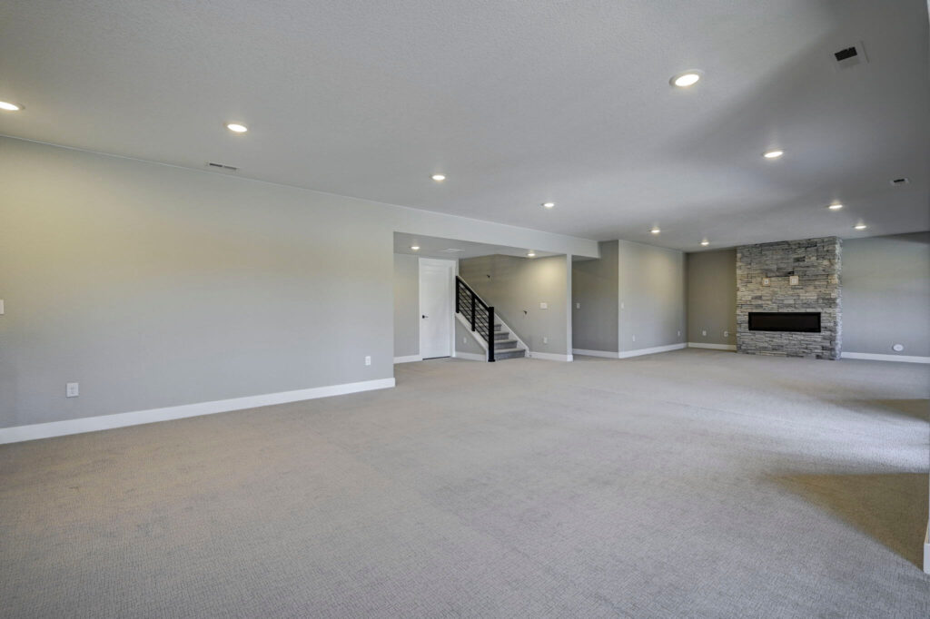 Large open carpeted basement room with recessed lights, stairs and a stone fireplace at far end.
