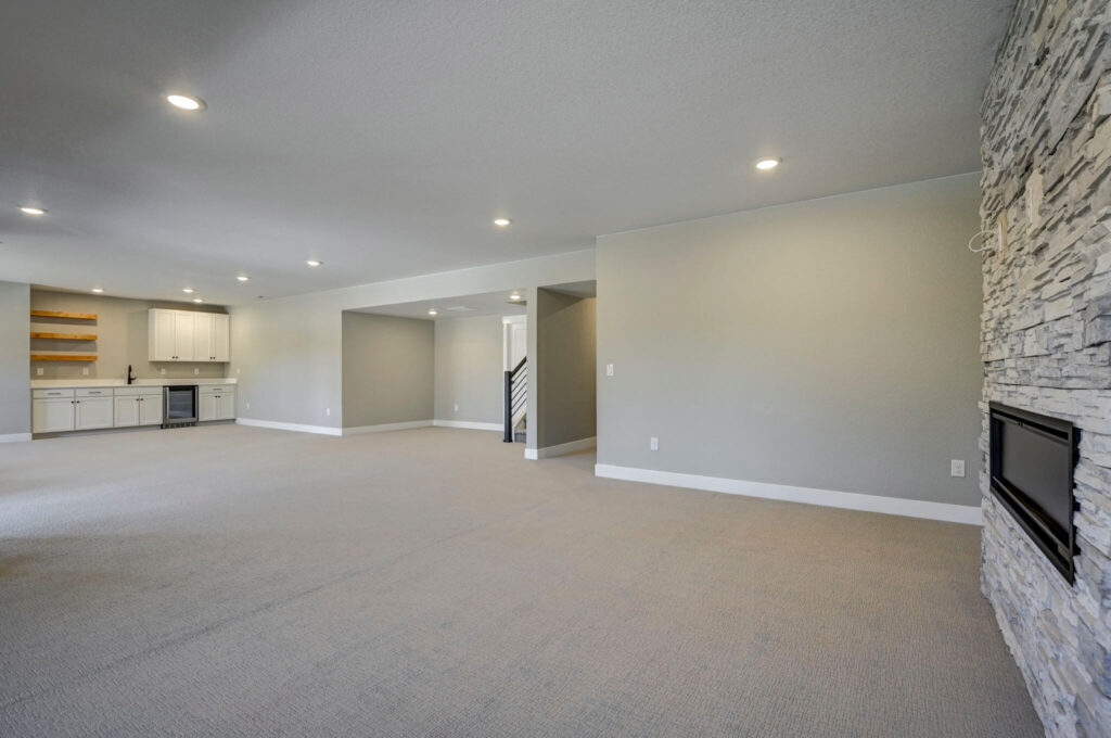 Open-plan basement view looking toward a wet bar and stone fireplace edge.