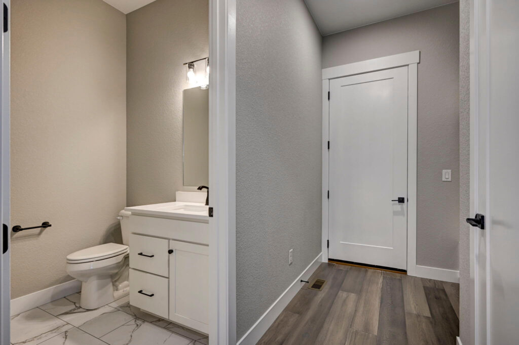 Small bathroom with white vanity, toilet, tiled floor and gray walls viewed from a hallway.