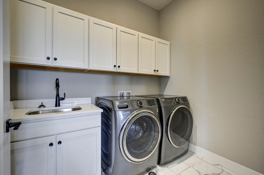 Laundry room with front-load washer and dryer, white cabinets, small sink, and gray walls.