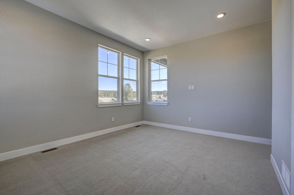 Empty carpeted bedroom with corner windows and neutral gray walls under recessed lighting.