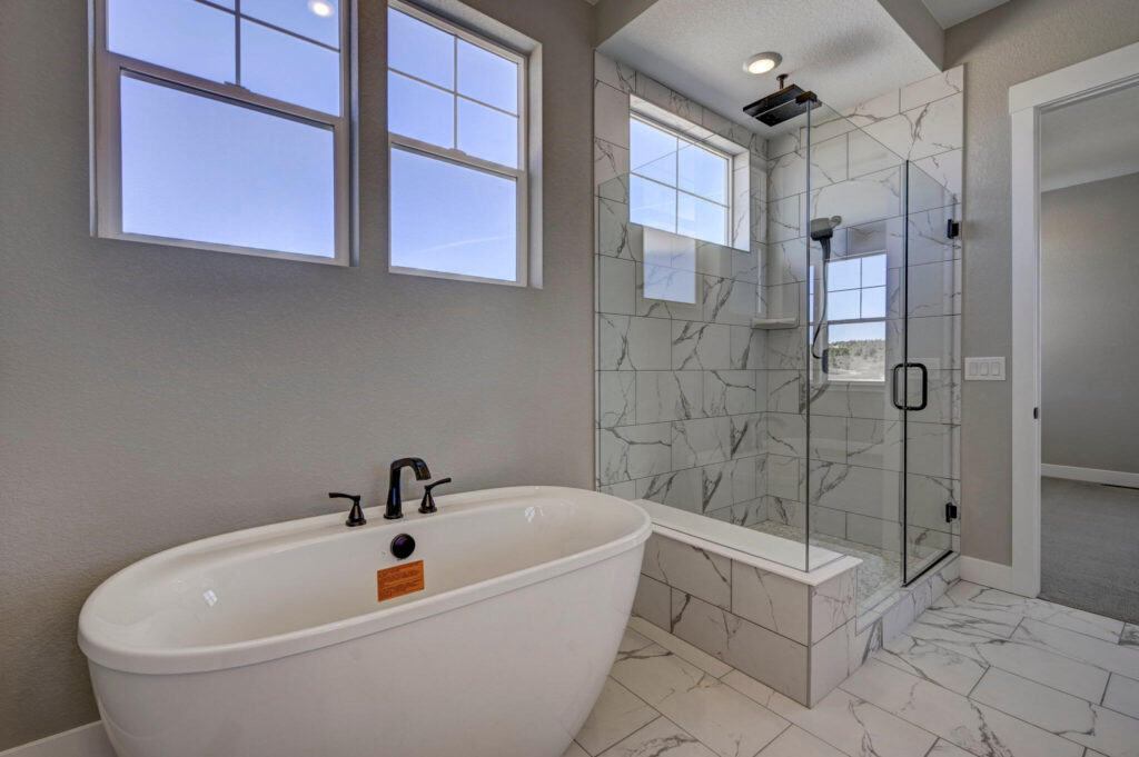 Freestanding tub beside a glass shower with large marble-effect tiles under two windows.