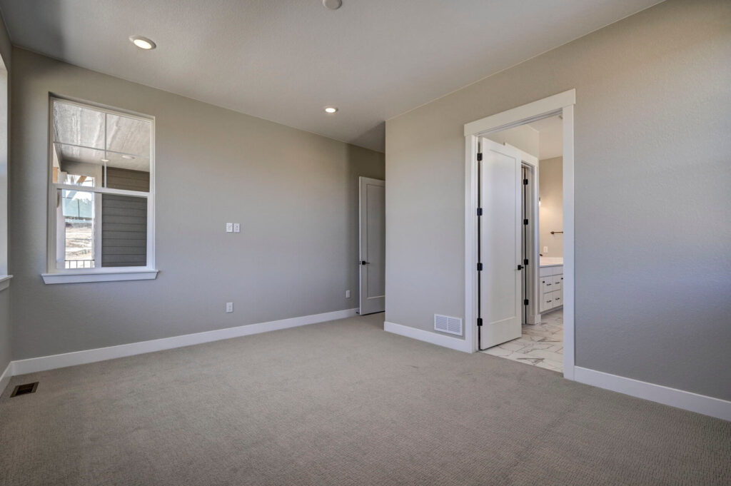 Empty carpeted room with light-gray walls, recessed lights, window, and doorway to a bathroom.