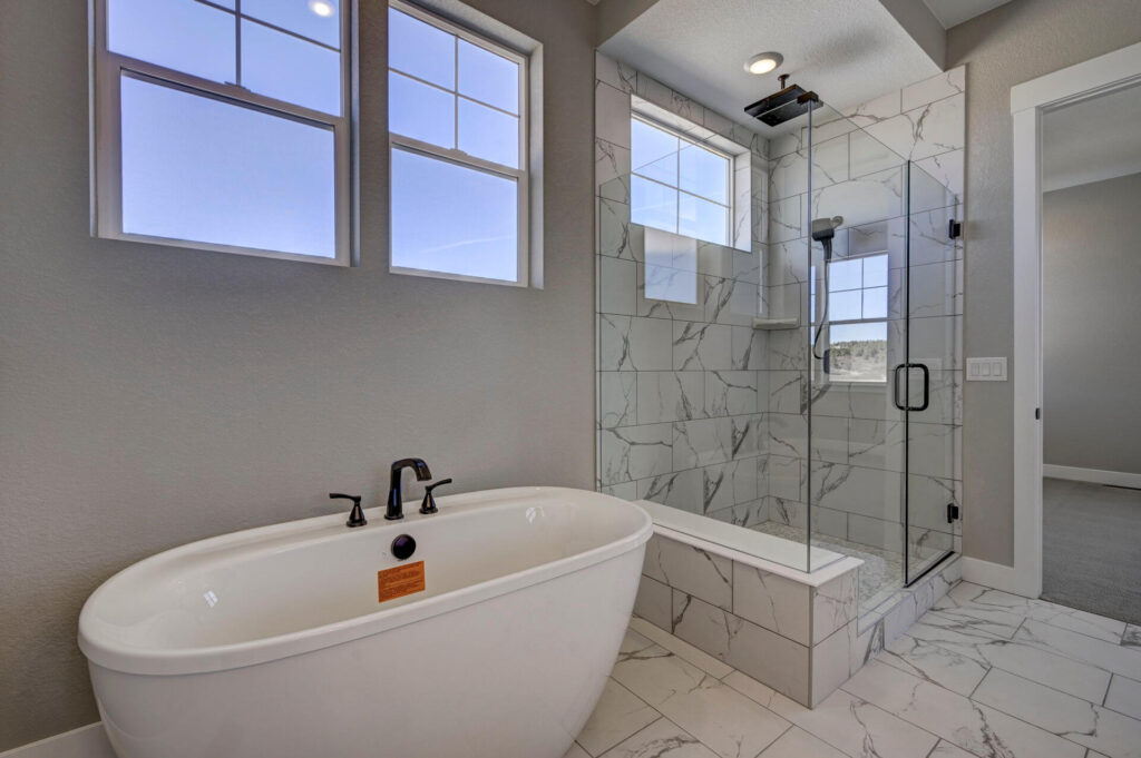Freestanding tub next to a glass shower with marble-pattern tile and two high windows.