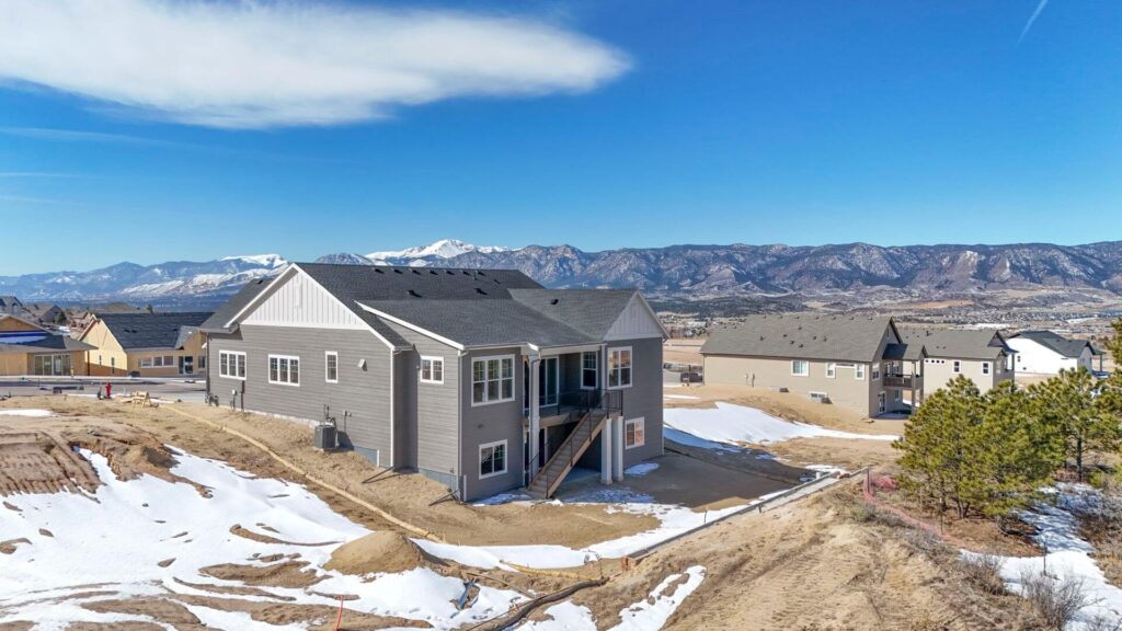 Gray two-level house on a hillside with snow patches and mountains beyond.