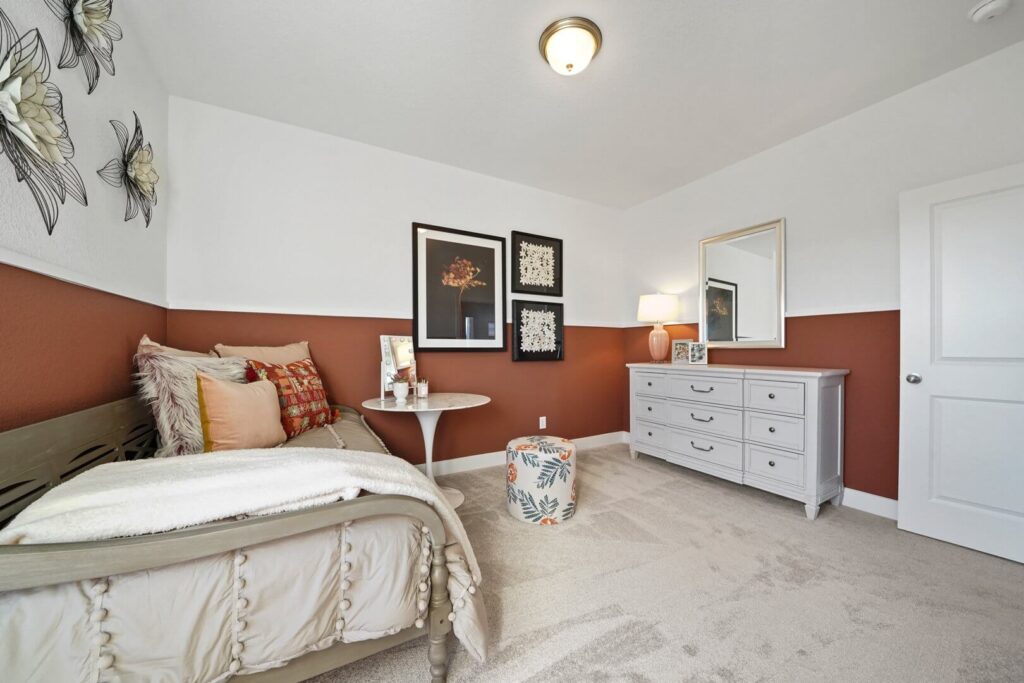 Wide angle of a bedroom with bed, dresser, framed wall art, and floral ottoman on carpet.