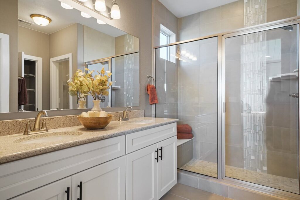 Bathroom vanity with dual sinks, large mirror, and glass-enclosed walk-in shower with tiled accent strip.