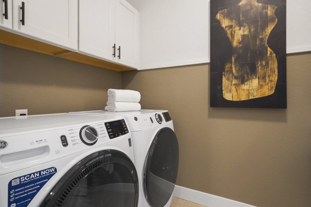 Laundry room with front-load washer and dryer under white cabinets and abstract wall art.