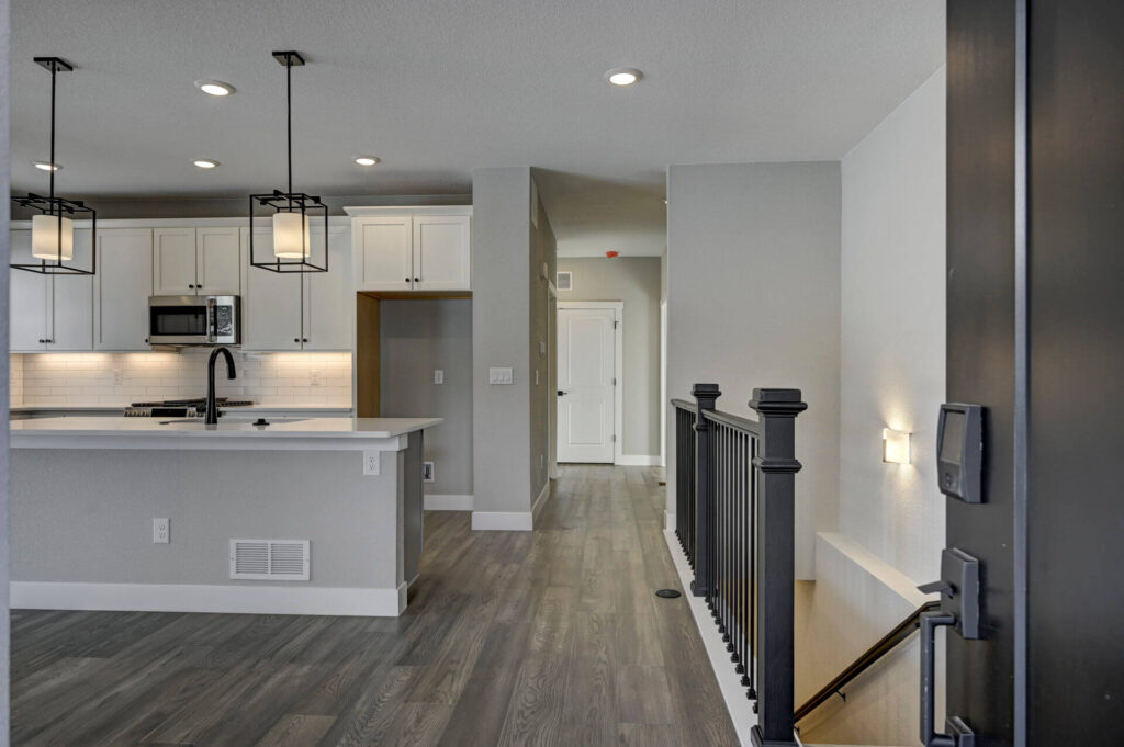 Open-plan kitchen and entry with island, pendant lights, gray floors and stair railing to the right.