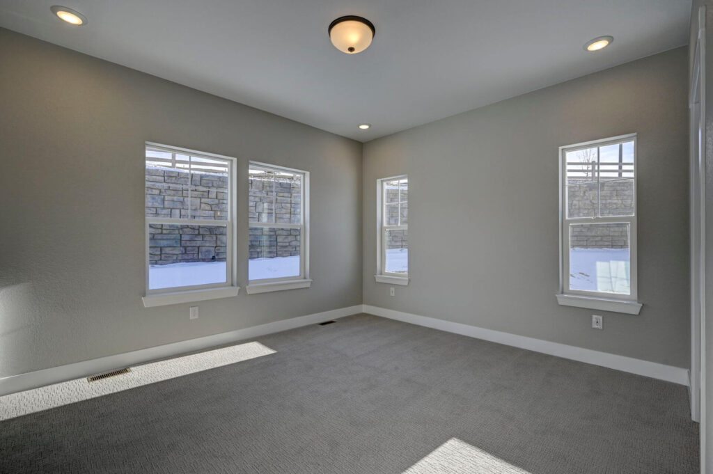 Empty carpeted room with three windows and neutral gray walls receiving daylight.
