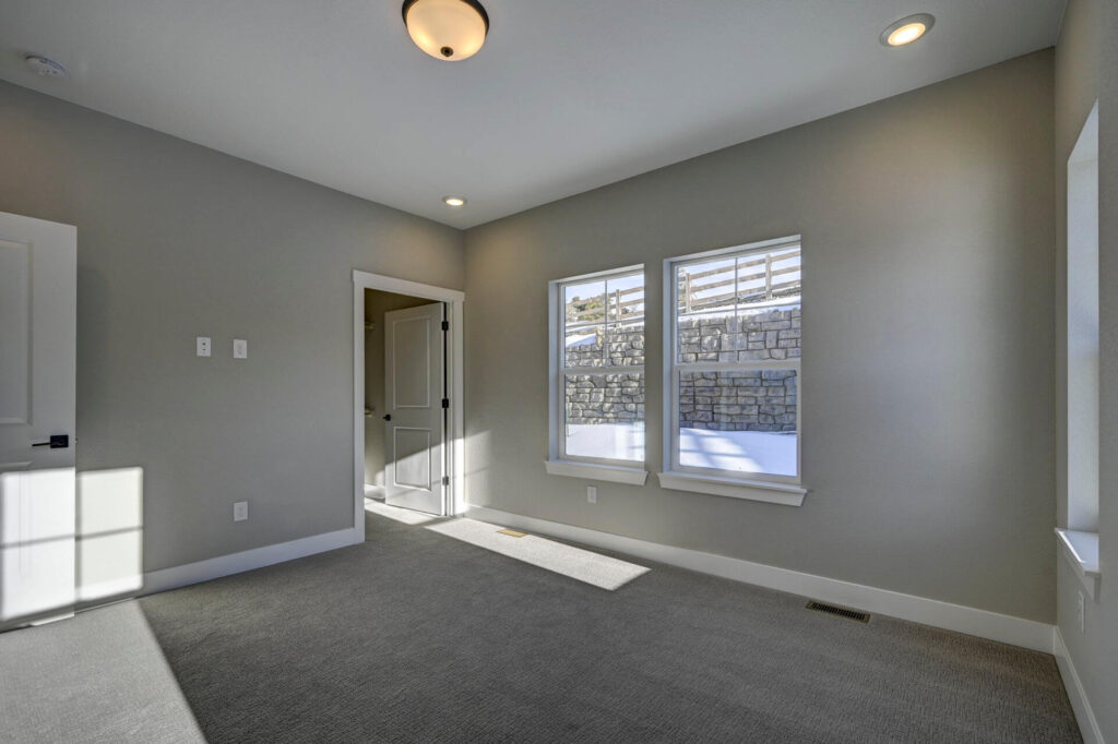 Another angle of a vacant carpeted room showing two windows, doorway, and ceiling lights.